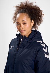 Kvinde med krøllet hår iført en mørkeblå hættetrøje med lynlås og hvide chevronmønstre på skulderen.