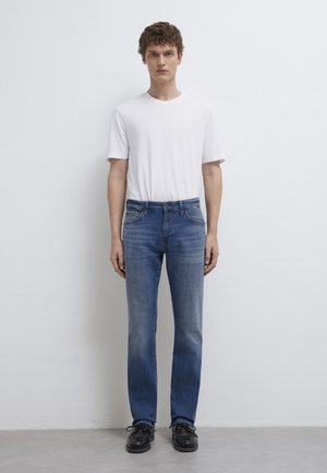 Junger Mann mit lockigem Haar, der ein schlichtes weißes T-Shirt, blaue Jeans und schwarze Schuhe trägt und vor einer weißen Wand steht.