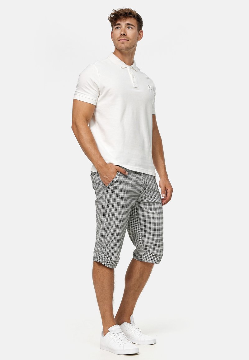 Weißes Polo-Shirt mit kurzen Ärmeln, graue karierte Shorts und weiße Sneaker. Das Outfit hat ein dezentes Logo auf dem Polo und umgeschlagene Shorts-Ärmel.
