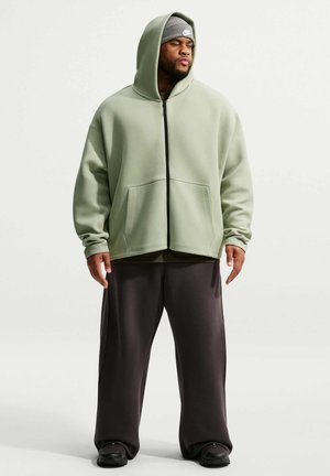 Man wearing sage green zip-up hoodie, gray beanie, loose black pants, and black sneakers standing against a plain light background.