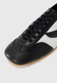 Chaussure de sport noire et blanche avec une tige en cuir texturé, des lacets plats noirs, une semelle en gomme et des coutures décoratives le long de l'avant-pied.