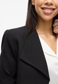 Černý blejzr se strukturovaným límcem, vyrobený z hladké tkaniny. Nosený přes bílý top, ukazující čistý design a moderní siluetu.