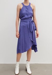Purple midi dress with a textured wave pattern, halter neckline, and a tied waist, featuring an asymmetrical hem. Worn with white ankle boots.