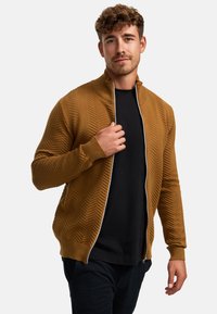 Brun zip-up jacka med texturerat herringbone-mönster, hög krage och ribbade mudd. Buren över en svart t-shirt. Slät tygfinish.