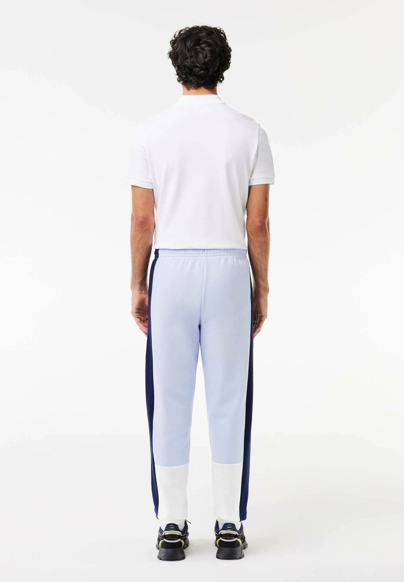 Homme portant un polo blanc à manches courtes, un pantalon de survêtement bleu clair avec des panneaux latéraux bleu marine et blancs, et des baskets noires, se tenant de dos sur un fond blanc.