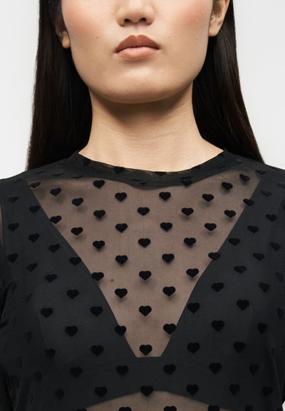 Γυναίκα που φοράει μαύρο διάφανο τοπ με ανάγλυφα βελούδινα μοτίβα σε σχήμα καρδιάς και βαθύ V-σχήματος ντεκολτέ.