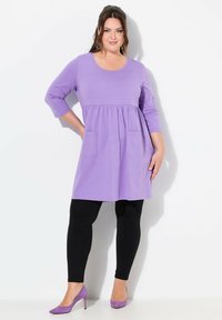 Lila tunikaklänning med rund halsringning, trekvartsärmar, samlad midja och två framfickor, i kombination med svarta leggings och lila klackar.