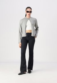 Jaqueta cinza curta com decote redondo, usada sobre um top cropped branco. Combinada com jeans pretos à boca de sino e sapatos pretos. A modelo usa óculos de sol.