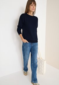 Marineblå sweatshirt med ribbede mansjetter og bund, parret med lyseblå denimjeans og beige sneakers. En kviltet off-white tote bag bæres.