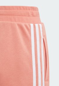 Joggers de color melocotón con una textura suave, que presentan tres franjas verticales blancas a lo largo del costado y un bolsillo insertado en el lado derecho.