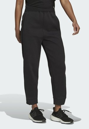 Persoon die zwarte joggerbroek met omgeslagen pijpen en zwart-witte sportschoenen draagt, staand tegen een eenvoudige lichte achtergrond.