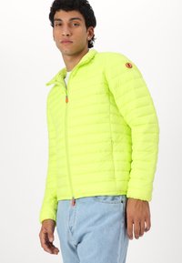 Bright neon yellow puffer jacket with a zip collar, featuring horizontal quilted sections, one sleeve logo, and an orange zipper pull detail.