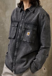 Tmavě šedá džínsová košile s knoflíkovým zapínáním, dvěma předními kapsami a jemně vybledlou texturou. Má malou nášivku s logem Carhartt.