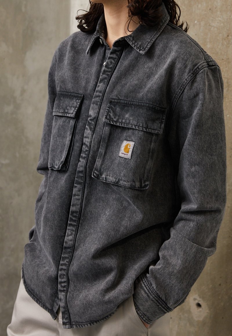 Donkergrijze spijkerblouse met knopenluiting, twee voorkruiszakken en een subtiele vervaagde textuur. Voorzien van een klein Carhartt logo label.