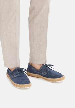 Piedi che indossano scarpe espadrillas blu con lacci, suole intrecciate beige e pantaloni beige chiaro su sfondo bianco.