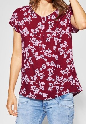 Vrouw draagt een bordeauxrode blouse met korte mouwen en lichtroze bloemenpatronen, gecombineerd met lichtblauwe spijkerbroek.