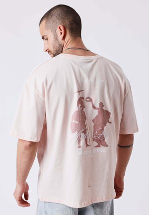 Homme aux cheveux courts portant un T-shirt oversized rose pâle avec un guerrier grec ancien et une figure échangeant une couronne de laurier dans le dos.
