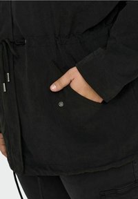 Svart jacka med dragsko i midjan, som har en sidficka, textilierad väv och synliga sömnadsdetaljer. Tyget har en matt yta.