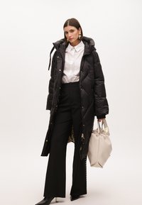Černý prošívaný dlouhý puffer kabát s kapucí. Bílá košile s knoflíky, černé pruhované kalhoty a béžová kabelka. Hladký povrch, zlaté akcenty.