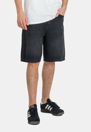 Male wearing black denim shorts, white t-shirt, black and white Adidas sneakers, one hand in pocket, standing against a plain background.