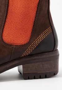 Ruskea nahkasaapas nilkkamalli, jossa on oranssi kudottu kangasosa, kontrastivärinen keltainen ommel, teksturoitu pohja ja lohkarekorko.