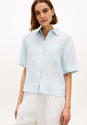 Frau trägt ein hellblaues, kurzärmliges Hemd mit Knöpfen und einer Brusttasche sowie weiße Hose vor einem einfarbigen Hintergrund.