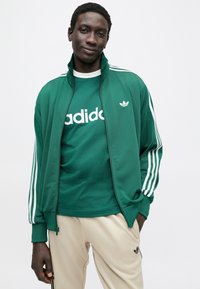 Grønn treningsjakke med glidelås og hvite striper på ermene, satt sammen med en grønn t-skjorte med fet hvit tekst, og beige bukser.