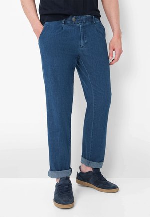 Uomo con jeans in denim blu risvoltati e sneaker blu navy con suole marrone chiaro, in piedi con le mani nelle tasche.