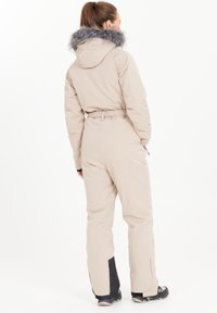 Beige jumpsuit med en vadderad design, med en krage av fuskpäls i grått, justerbar midja och svarta detaljer vid anklarna och fickorna.