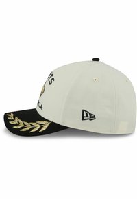 White cap with a black visor, gold laurel embroidery, and decorative text. Features a logo on the side. Cotton material with structured design.