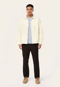 Crème-kleurige button-up jas met twee frontzakken over een lichtblauw shirt, gecombineerd met zwarte broeken en tan schoenen.