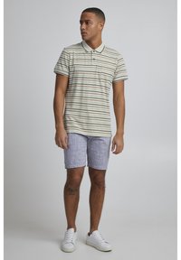 Stripet polo skjorte i beige med brune og marineblå striper, sammen med lysegrå shorts. Har krage og brystlomme. Hvite sneakers.