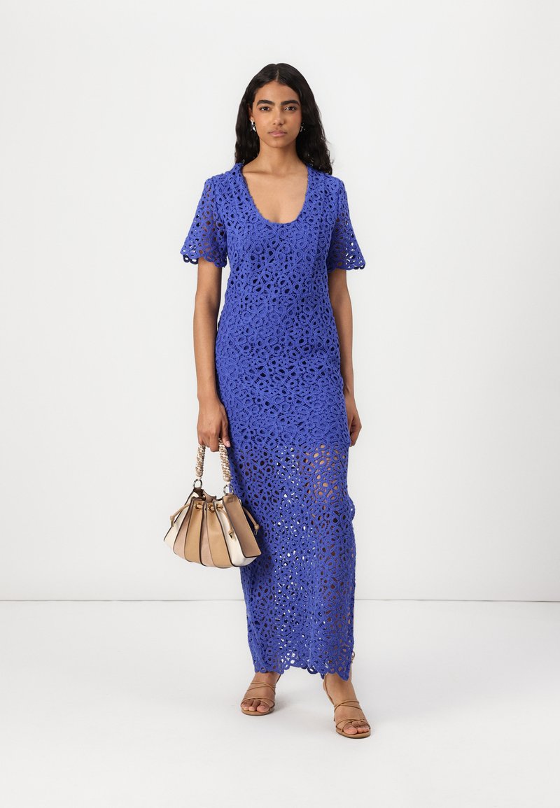 Maxikleid aus blauer Spitze mit kurzen Ärmeln, das einen Rundhalsausschnitt und ein aufwendiges florales Muster aufweist, kombiniert mit einer hellbraunen Handtasche und Sandalen.