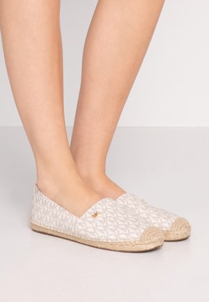 Pies con zapatos espadrilles beige sin cordones con suelas tejidas y parte superior de tela con un diseño de logo repetido.