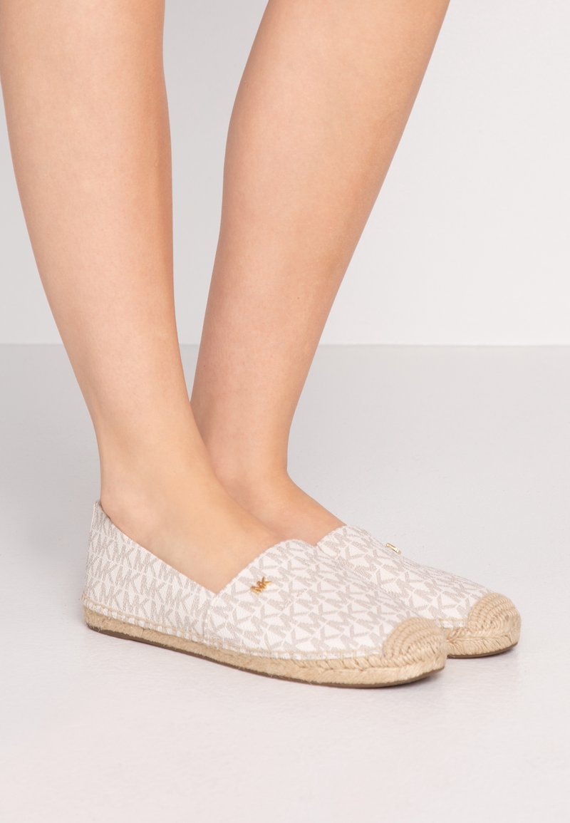 Pieds portant des espadrilles beiges sans lacets avec semelles tissées et dessus en tissu à motifs avec un logo répété.