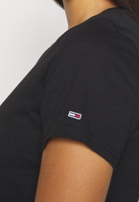 Černé bavlněné tričko s přiléhavým střihem. Mezi nápadné detaily patří malá nášivka s logem na rukávu, která obsahuje červené, bílé a modré barvy.