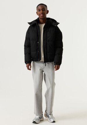 Tienerjongen staat en glimlacht, draagt een zwarte pufferjas, lichtgrijze jeans, een beige shirt en grijze hardloopschoenen tegen een effen achtergrond.