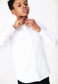 Camisa blanca de botones con cuello puntiagudo, de textura suave y mangas largas con puños abotonados, usada por una persona que ajusta el cuello.