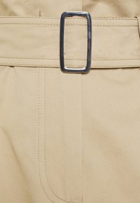 Ceinture en tissu beige avec une boucle rectangulaire en argent, présentant des coutures nettes et une texture lisse. Gros plan sur les détails de la ceinture et de la couture du pantalon.