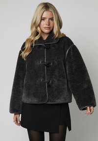 Femme blonde portant un manteau en peluche gris foncé avec des boutons en bois sur une jupe noire et des collants noirs, se tenant devant un fond uni.