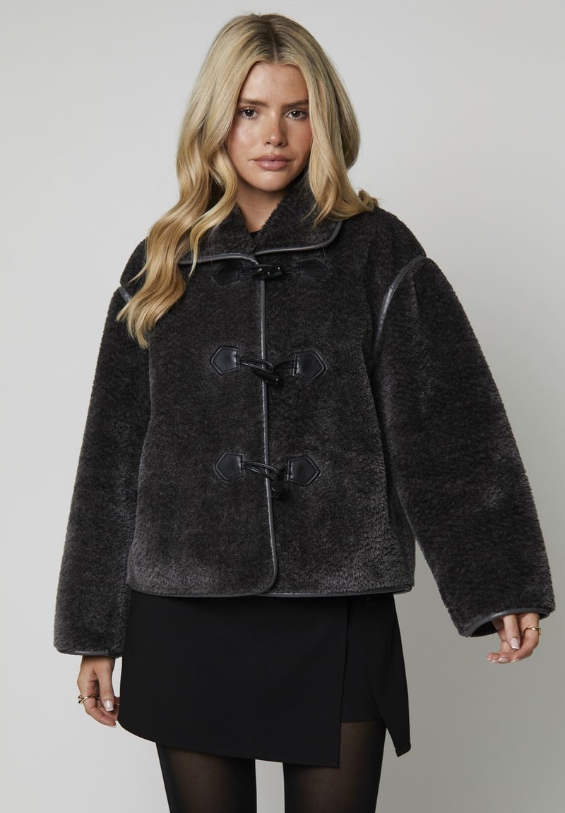 Femme blonde portant un manteau en peluche gris foncé avec des boutons en bois sur une jupe noire et des collants noirs, se tenant devant un fond uni.