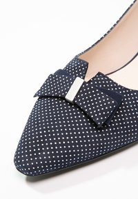 Zapato plano de lunares en azul marino con acabado de ante texturizado, puntera puntiaguda y un lazo decorativo con detalles en metal de tono plateado.
