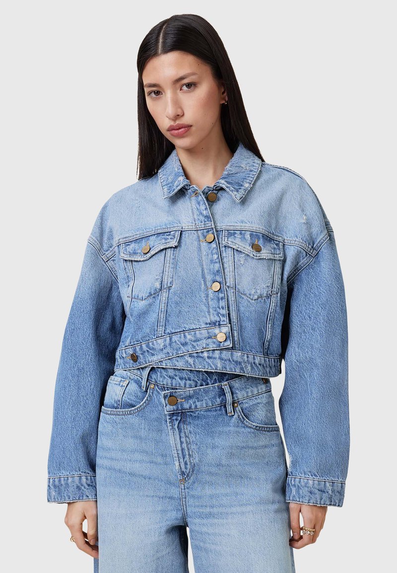 Chaqueta de mezclilla azul claro, recortada, con mangas largas, bolsillos frontales y detalles de botones de latón. Presenta una textura deslavada y un diseño de cuello clásico.