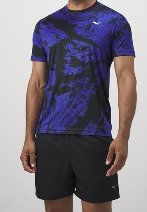 Homme portant un t-shirt de sport Puma violet et noir à motif abstrait et un short Puma noir sur un fond uni.