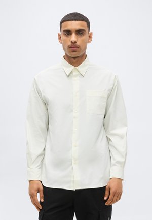 Uomo con capelli corti e baffi che indossa una camicia bianca con bottoni e taschino sul petto e pantaloni neri, in piedi davanti a uno sfondo semplice.