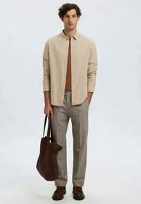 Camisa bege de botões por cima de uma blusa castanha, calças cinzentas, sapatos castanhos e uma mala tote castanha-escura. Design simples com um ajuste relaxado.