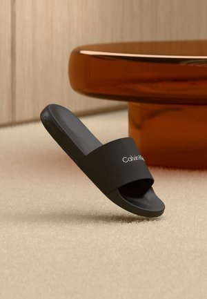 Sort Calvin Klein-sandalslide svæver over beige tæppe nær buet bordben i ravfarve.
