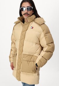 Beige dunjakke med kviltet design, høy krage, to framlommer og en liten logo-patch på brystet.