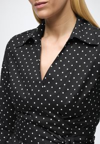 Zwarte wikkeljurk met witte polka dots, voorzien van een kraag en een gladde textuur. Het ontwerp heeft een gestrikte taille en een V-hals.