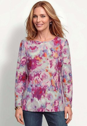 Frau mit schulterlangem blondem Haar, die ein farbenfrohes Langarmshirt mit pinken, violetten, orangen und blauen abstrakten Mustern und blaue Jeans trägt.